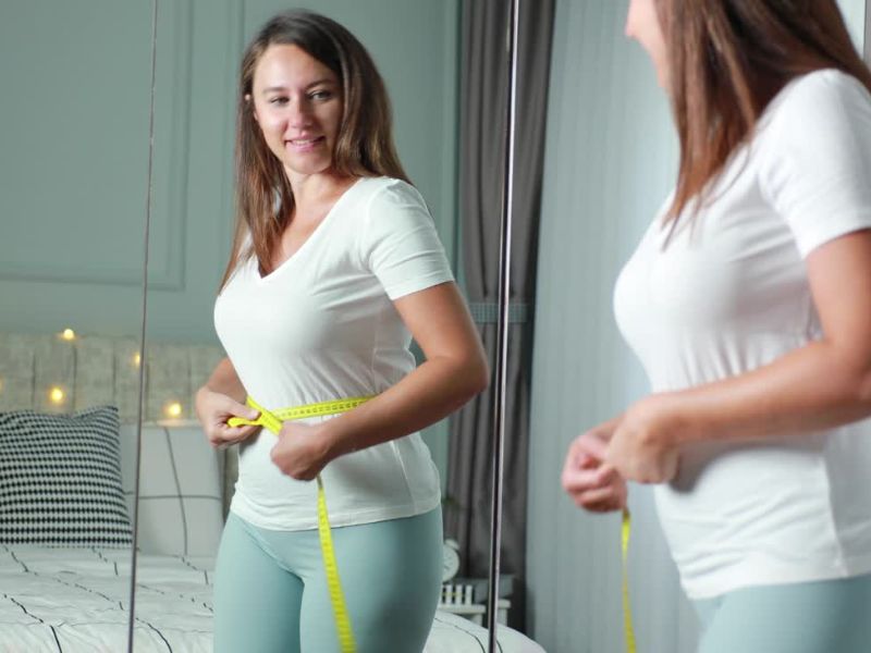 devojka koristi metar za merenje struka i zadovoljno se gleda u ogledalo